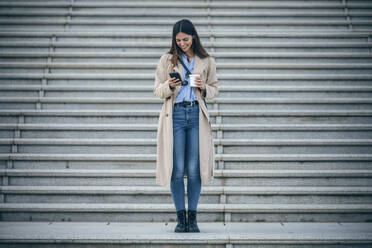 Glückliche Frau mit Kaffeetasse und Smartphone vor einer Treppe - JSRF02647