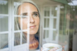 Reife Frau mit Teetasse schaut durch das Fenster - KNSF09835