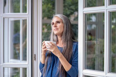 Thoughtful mature woman holding tea cup in doorway - KNSF09846