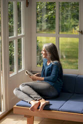 Smiling woman sitting with book on sofa at home - KNSF09852