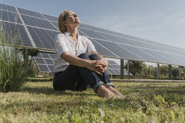Mature engineer sitting on grass with eyes closed in front of solar panels - OSF02153