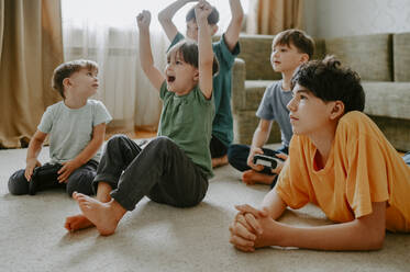 Glücklicher Junge spielt Videospiel mit Freunden, die zu Hause auf dem Teppich jubeln - ANAF02126