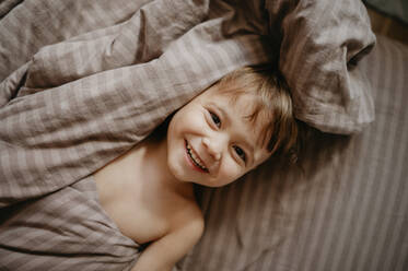 Lächelnder Junge auf dem Bett liegend mit Decke zu Hause - ANAF02141