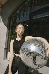 Glückliche Frau hält Discokugel an einem sonnigen Tag - YBF00249