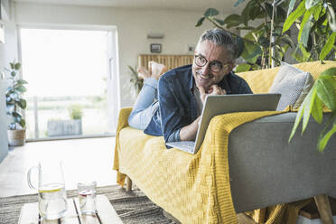 Nachdenklicher Freiberufler mit Laptop, der zu Hause auf dem Sofa liegt - UUF30263