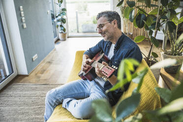 Smiling mature man playing guitar sitting on sofa at home - UUF30268