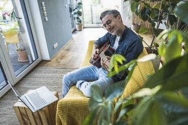 Gitarre spielender Mann auf dem Sofa sitzend mit Laptop zu Hause - UUF30269