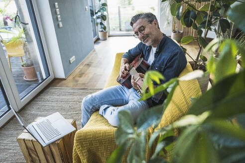 Gitarre spielender Mann auf dem Sofa sitzend mit Laptop zu Hause - UUF30269