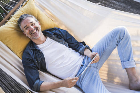 Happy mature man with tablet PC relaxing in hammock - UUF30277