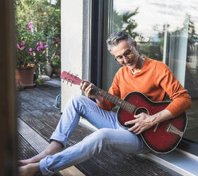 Smiling man playing guitar sitting on porch seen through glass - UUF30284