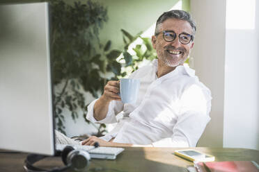 Happy businessman holding coffee cup at home office - UUF30294