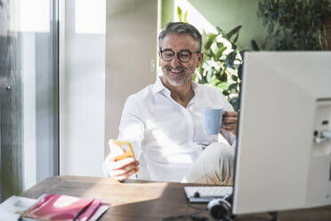 Lächelnder Freiberufler, der ein Smartphone benutzt und eine Kaffeetasse im Büro zu Hause hält - UUF30295