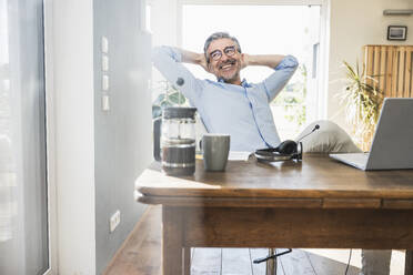 Happy businessman with hands behind head sitting at desk in home office - UUF30317