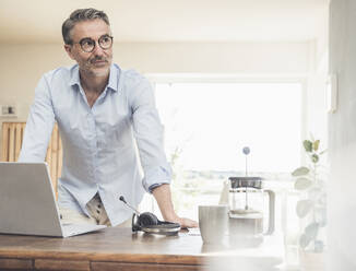 Thoughtful freelancer leaning on table with laptop at home office - UUF30320