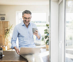 Smiling businessman with laptop holding cup sitting on desk in home office - UUF30323