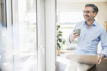 Happy businessman with laptop and cup sitting at home office - UUF30324