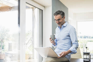 Mature businessman with laptop and cup sitting on desk at home office - UUF30325