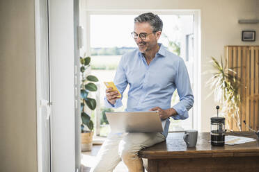 Lächelnder Geschäftsmann mit Laptop und Smartphone auf dem Schreibtisch im Heimbüro - UUF30326