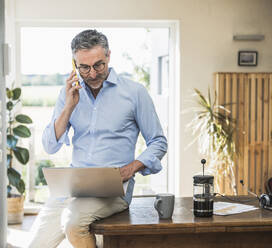 Geschäftsmann, der mit seinem Smartphone telefoniert und einen Laptop im Home Office benutzt - UUF30327