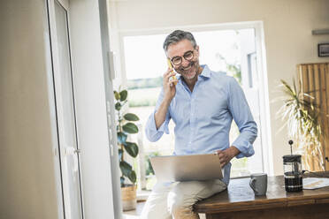 Happy businessman talking on mobile phone sitting on desk at home office - UUF30328