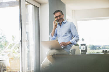 Smiling businessman talking over smart phone sitting on desk at home office - UUF30329