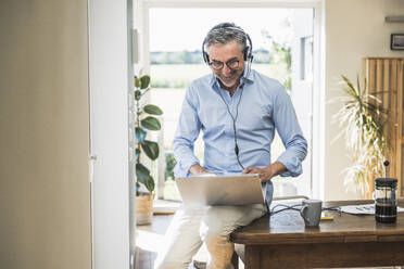 Lächelnder Geschäftsmann mit Headset bei der Arbeit am Laptop im Home Office - UUF30333