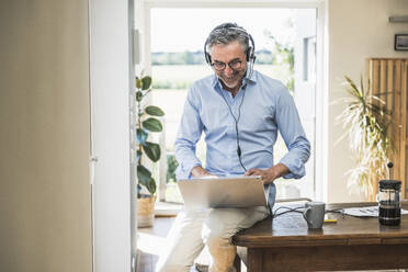 Smiling businessman with headset working on laptop at home office - UUF30333