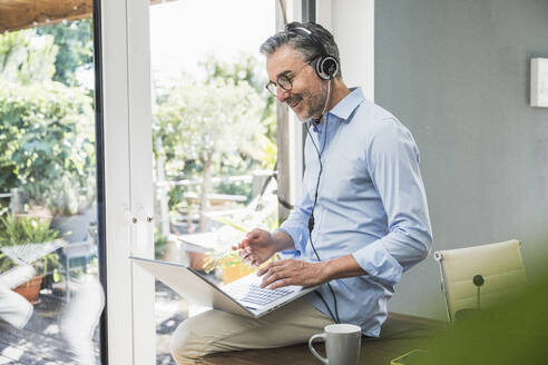 Lächelnder Geschäftsmann mit Laptop und Headset im Heimbüro - UUF30336