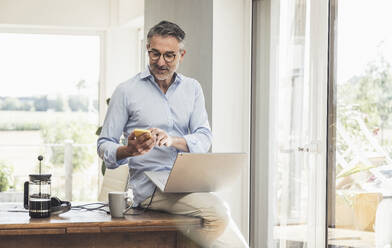 Businessman using smart phone sitting on desk at home office - UUF30337