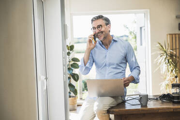 Smiling freelancer talking on mobile phone at home office - UUF30338