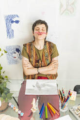 Künstlerin hält Bleistift zwischen Nase und Lippe am Tisch - MGRF01050