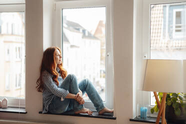 Thoughtful redhead woman sitting on window sill - KNSF09870