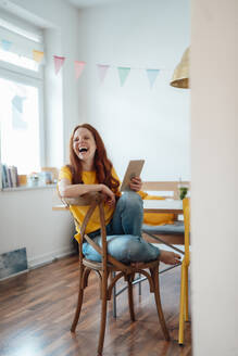 Fröhliche Frau lachend und sitzend mit Tablet-PC auf Stuhl zu Hause - KNSF09872