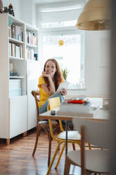 Happy woman sitting with hand on chin at table - KNSF09875