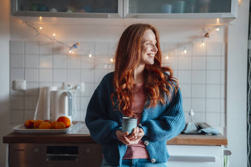 Happy redhead woman standing with coffee cup at home - KNSF09879