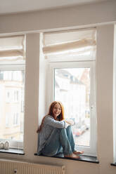 Happy woman sitting on window sill at home - KNSF09892