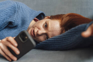 Frau liegt auf einem grauen Sofa und benutzt ein Smartphone - KNSF09893