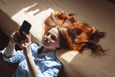 Smiling redhead woman using smart phone and leaning on bed at home - KNSF09904