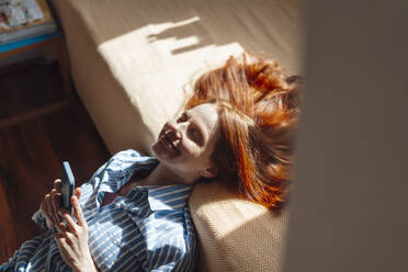 Smiling redhead woman with eyes closed leaning on bed at home - KNSF09905