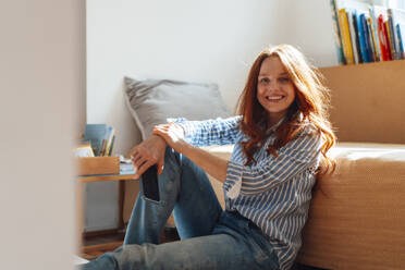 Happy redhead woman sitting with smart phone near bed at home - KNSF09908