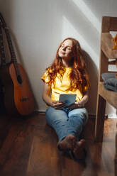 Smiling redhead woman sitting with eyes closed and tablet PC on floor - KNSF09916