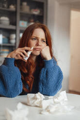 Traurige kranke Frau sitzt mit Medikamenten und Papiertüchern am Tisch - KNSF09922