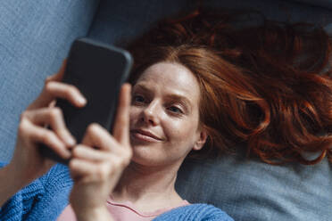 Smiling redhead woman using smart phone on sofa - KNSF09926