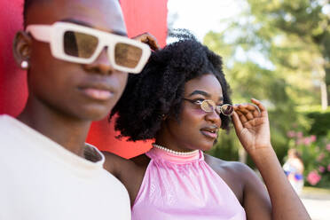 Porträt eines stilvollen schwarzen Teenagerpaares mit Afrofrisur und weißem Hemd mit trendiger Sonnenbrille, das vor einem roten Hintergrund und Bäumen in einem Park steht und wegschaut - ADSF47123