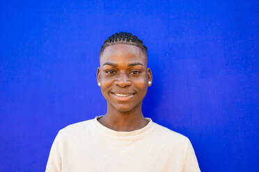 Portrait of confident African American teenage male in braids and white shirt standing against blue background while smiling and looking at camera - ADSF47124