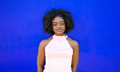 Portrait of African American young female in casual cloth with Afro hairdo and makeup standing against blue background and looking at camera - ADSF47128