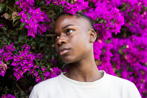 Portrait of serious African American teenage male in white shirt standing near blooming purple flowers in park while looking away - ADSF47130