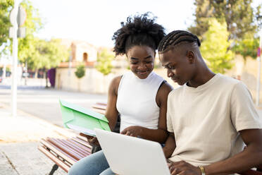 Smiling African American teenage couple in casual clothes sitting on wooden bench with cardboard and papers while typing on laptop in daylight - ADSF47134