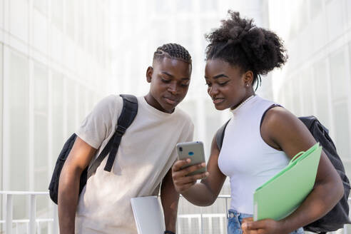 Focused African American teenage couple in casual clothes with backpacks browsing mobile phone while standing on street together in daylight - ADSF47137