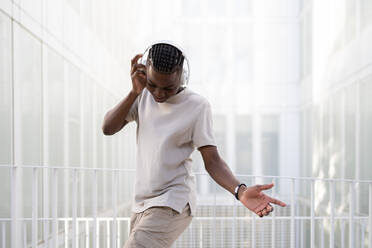 Side view of positive African American teenage male in casual clothes and headphones dancing while listening to music in wireless headphones on bright city banner - ADSF47140